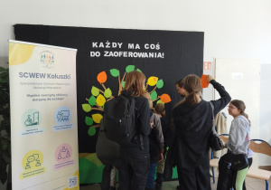 Młodzież i dorośli przywieszają swoje listki na drzewie, nad drzewem napis "Każdy ma coś do zaoferowania"